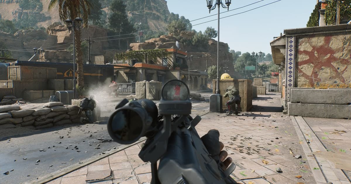 Ego-Perspektive in Delta Force: Ein Gewehr mit Rotpunktvisier und aufgesetztem Zielfernrohr zielt eine sonnige mediterrane Straße mit Sandsäcken entlang, vorne gehen zwei Gegner in Deckung