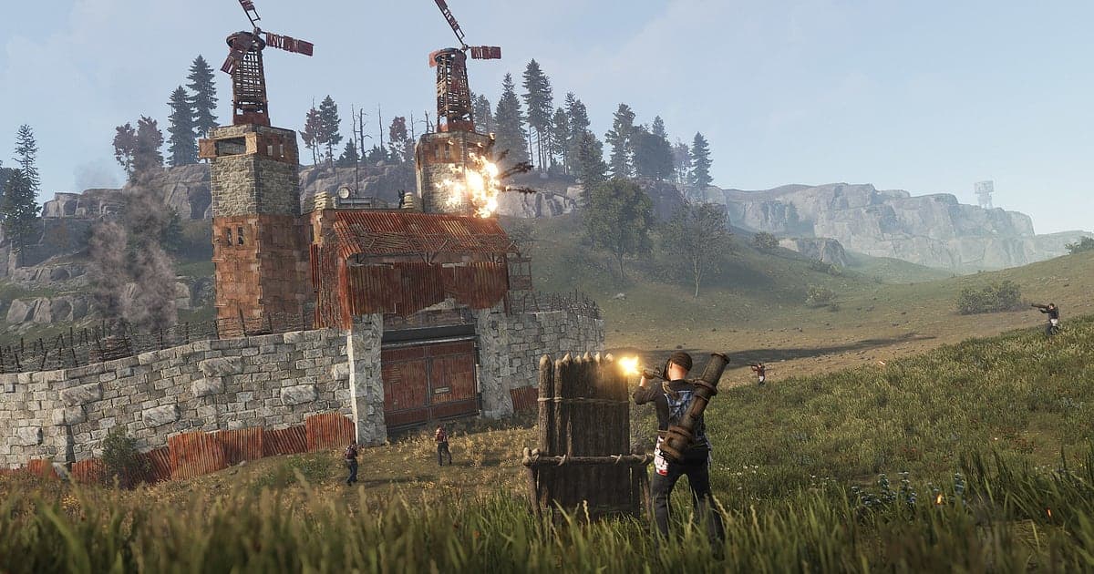 Eine Explosion im Obergeschoss einer Basis aus Stein und Blech in Rust, während unten im Feld ein Spieler mit Raketenwerfer hinter einer Holzbarrikade feuert