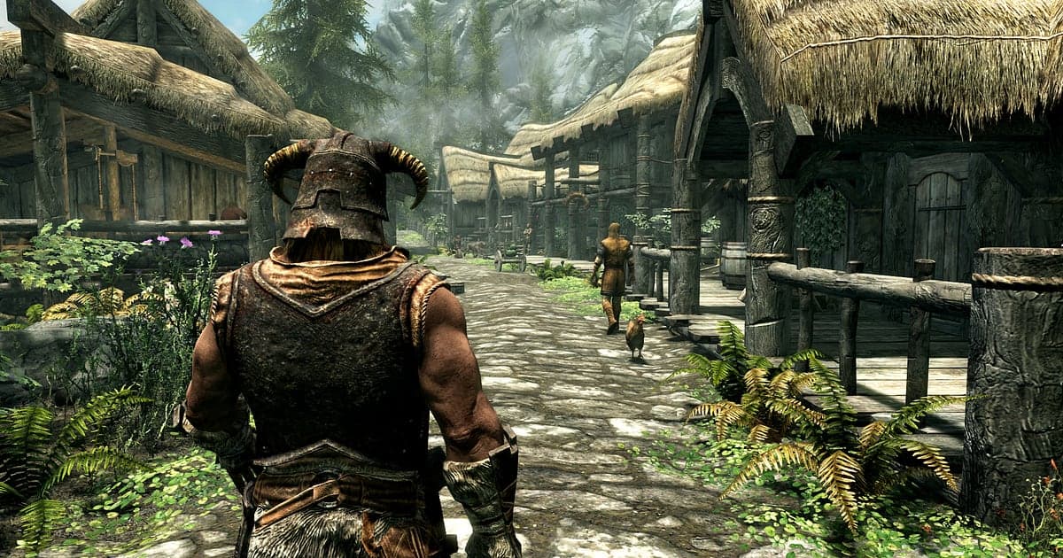 Szene aus Skyrim Special Edition, in der das Drachenblut mit Hörnerhelm einen steinernen Dorfweg zwischen strohgedeckten Fachwerkhäusern entlanggeht, in der Ferne verschneite Felsgipfel