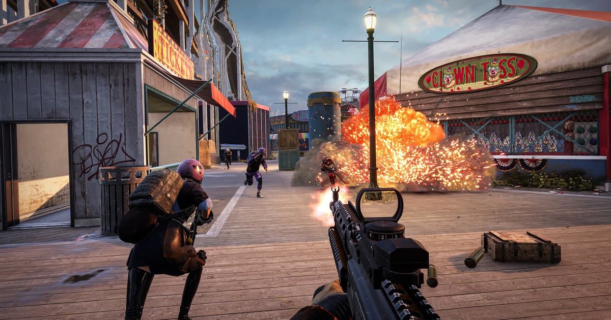 Visão em primeira pessoa de PUBG: BATTLEGROUNDS atirando com um fuzil com mira red dot num calçadão de parque de diversões, com uma explosão à frente, inimigos chegando correndo e um companheiro de squad agachado ao lado